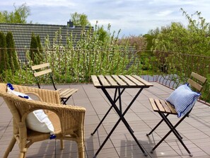 Eine Terrasse mit Tisch und zwei Stühlen, umgeben von einem Metallzaun und Pflanzen.