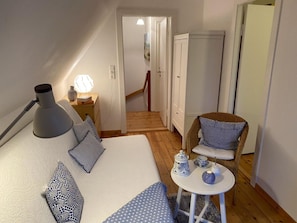 Ein Schlafzimmer mit Bett, Nachttischlampe, Kissen, einem Sessel, einem kleinen Tisch mit Teekanne und einer Tür zu einem anderen Raum.