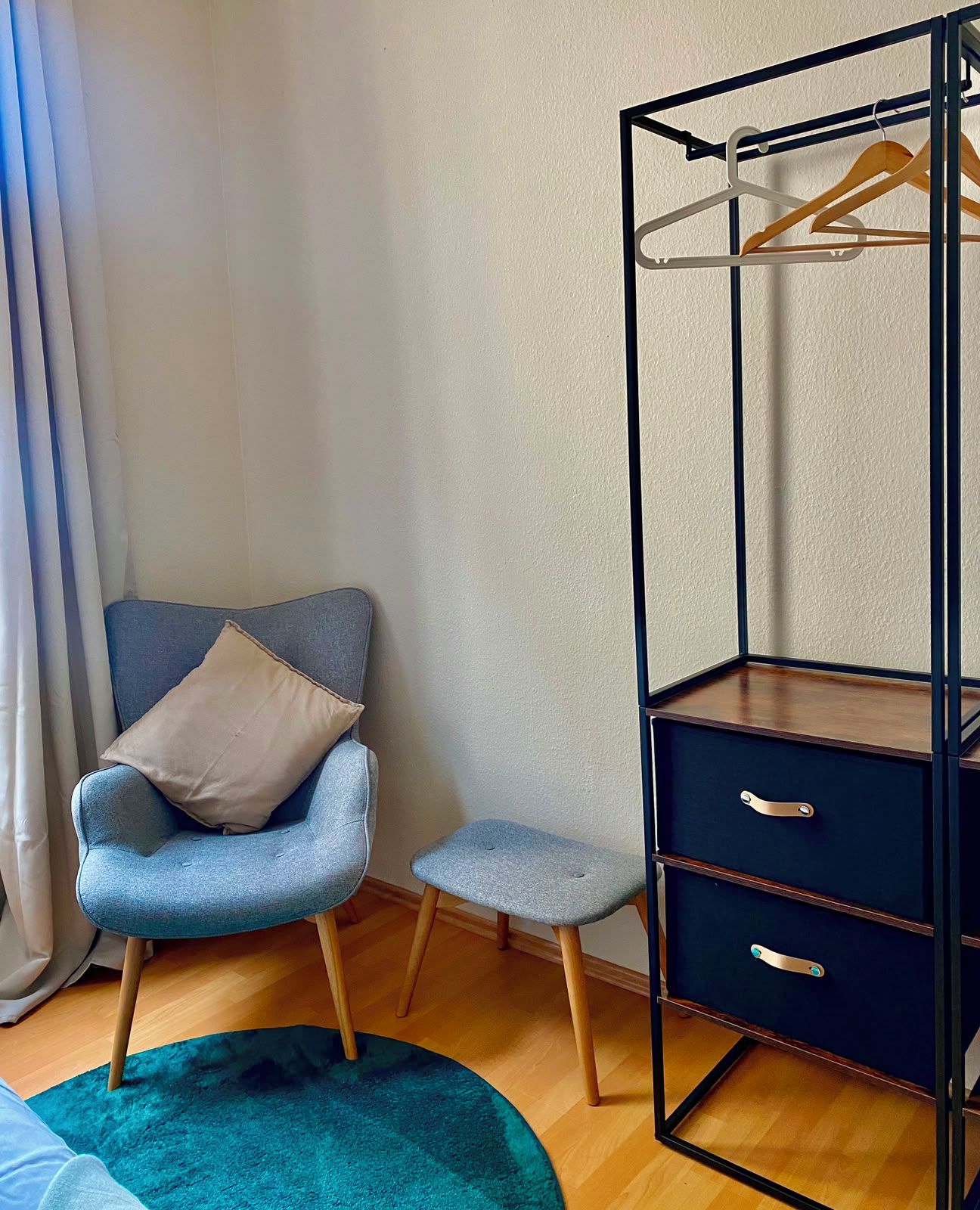 A room with a blue armchair, a small stool, a dark dresser with drawers, and a hanging clothes rack.