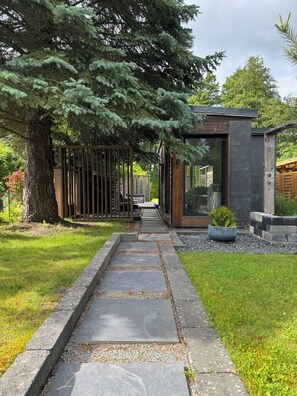 Uma trilha leva a um edifício moderno com porta de vidro, cercado por um jardim bem cuidado e uma grande árvore.