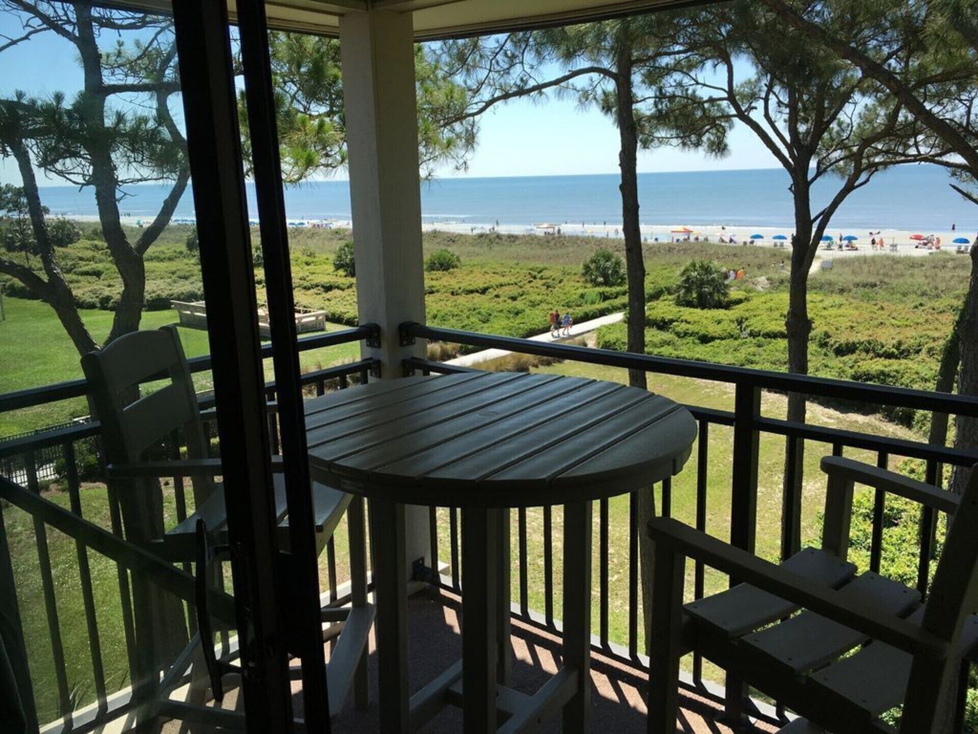 A balcony with a table and chairs, overlooking a beach with people and umbrellas.