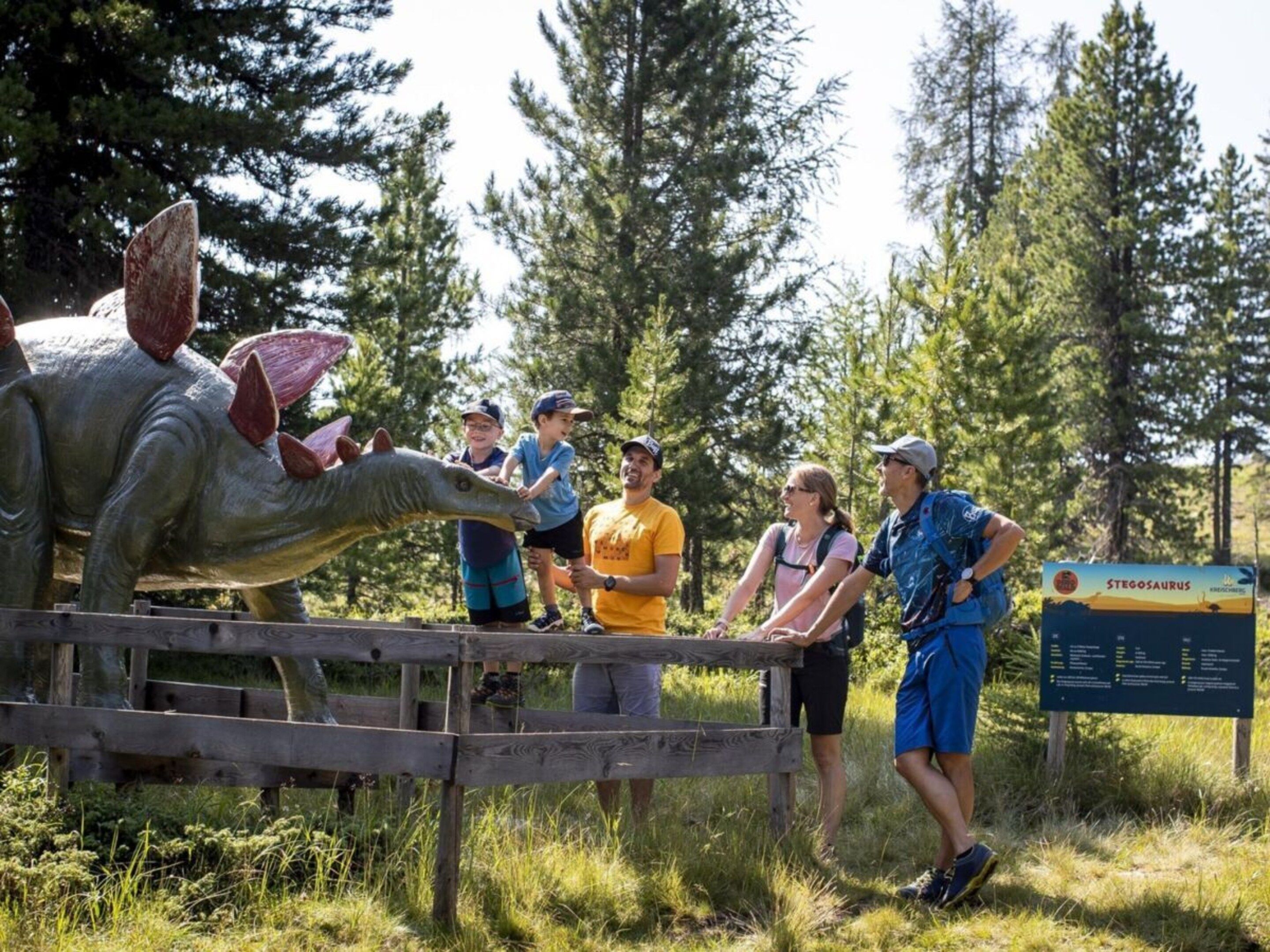 Un grupo de personas observa una exhibición de dinosaurios al aire libre, con un cartel de fondo.