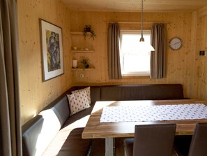 Ein gemütliches Zimmer mit Holzeinrichtung, einem Esstisch mit gemustertem Tischlauf, einer Couch mit Kissen sowie einer Wanduhr und einem Fenster mit Vorhängen.