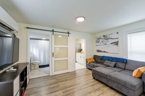 A modern living room with a grey sofa, a flat-screen TV, and a sliding door leading to a bathroom.