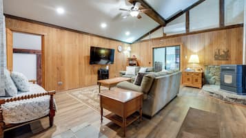 A cozy living room with a wooden floor, a sofa, a wooden coffee table, a TV mounted on the wall, and a stone fireplace.