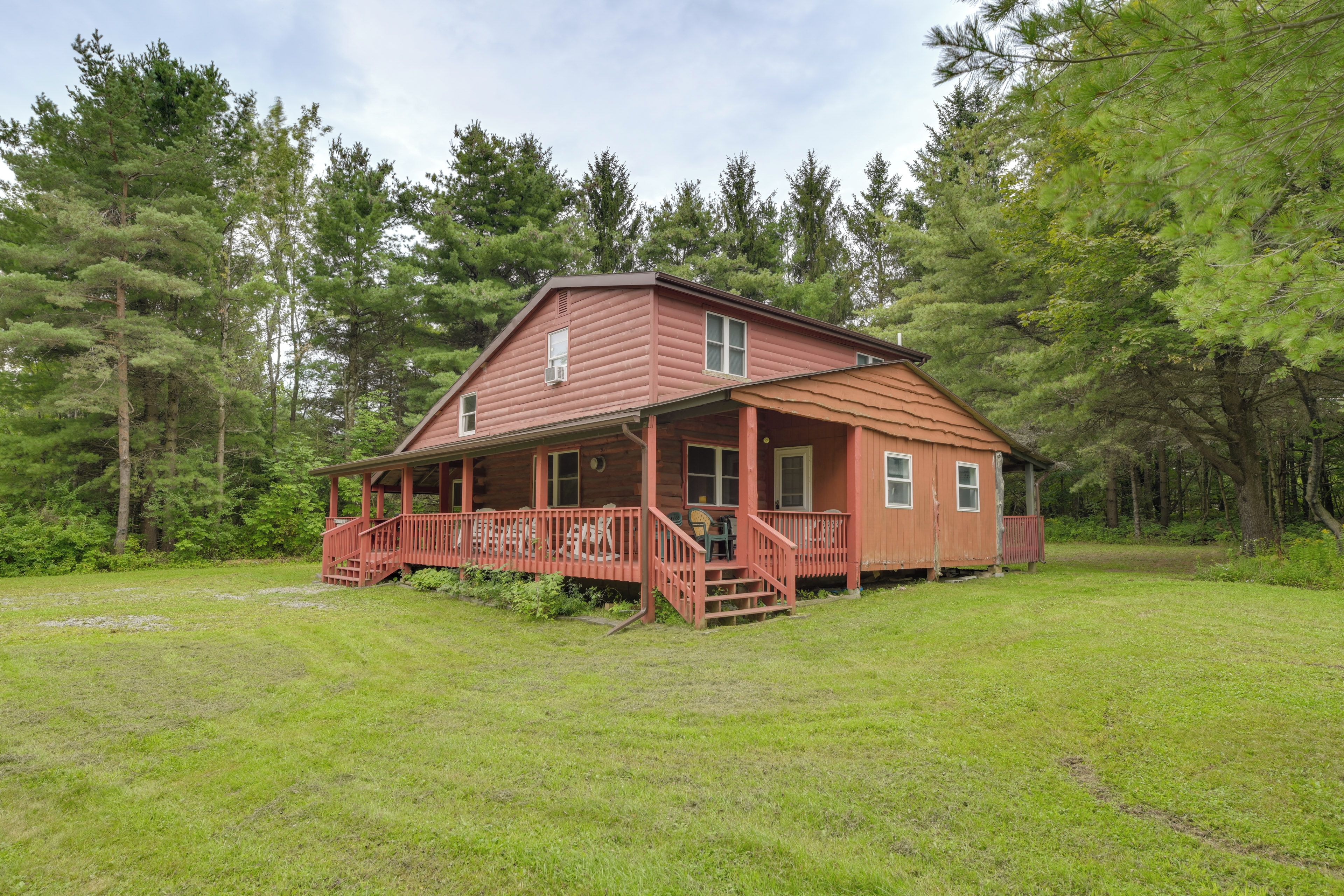 A two-story wooden cabin with a porch and a small deck, surrounded by a grassy field and dense forest.