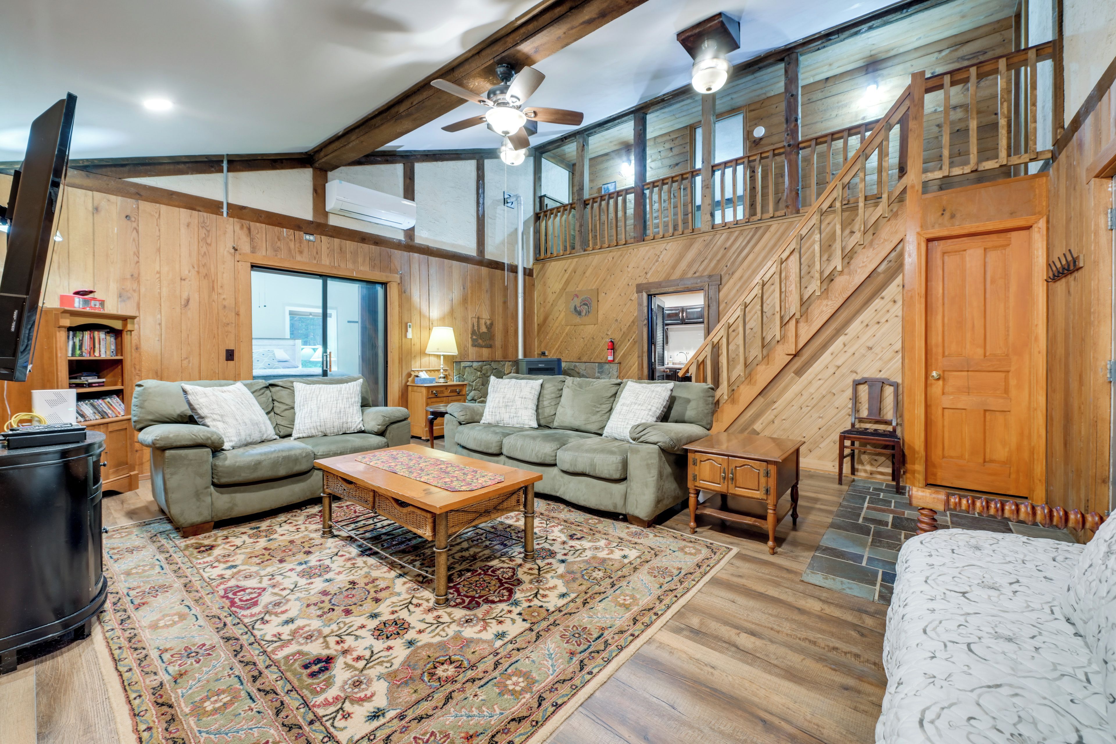 A spacious living room with a high ceiling, wooden beams, a staircase, and a large window.
