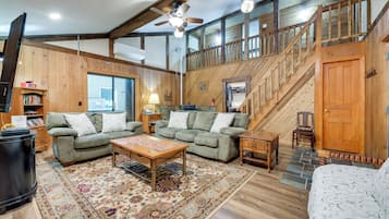 A spacious living room with a high ceiling, wooden beams, a staircase, and a large window.