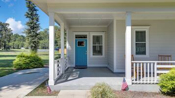 A white house with a blue door, a covered porch, and a small garden with an American flag.