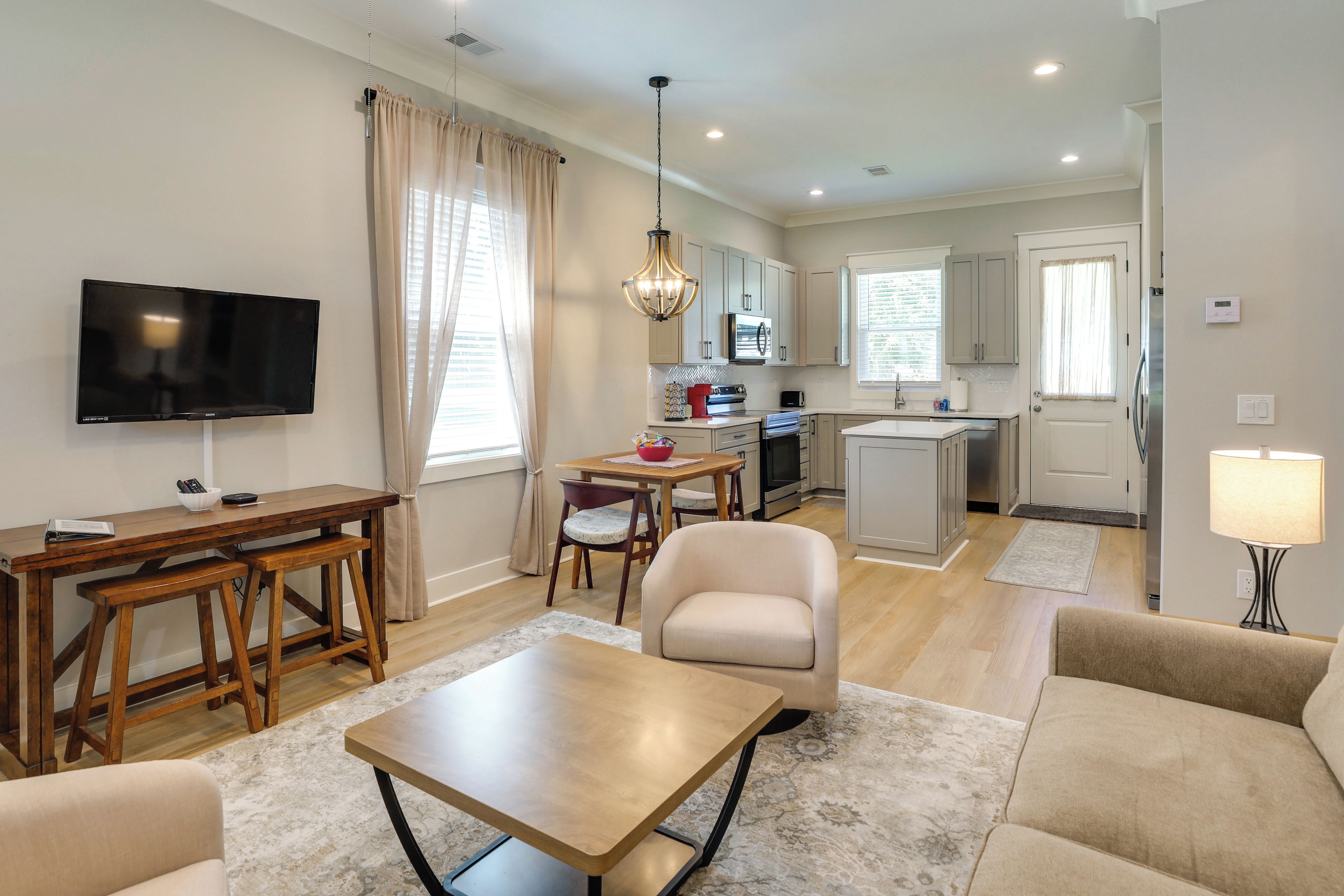 A modern living room with a flat-screen TV, wooden furniture, a dining area, and a kitchen with stainless steel appliances.