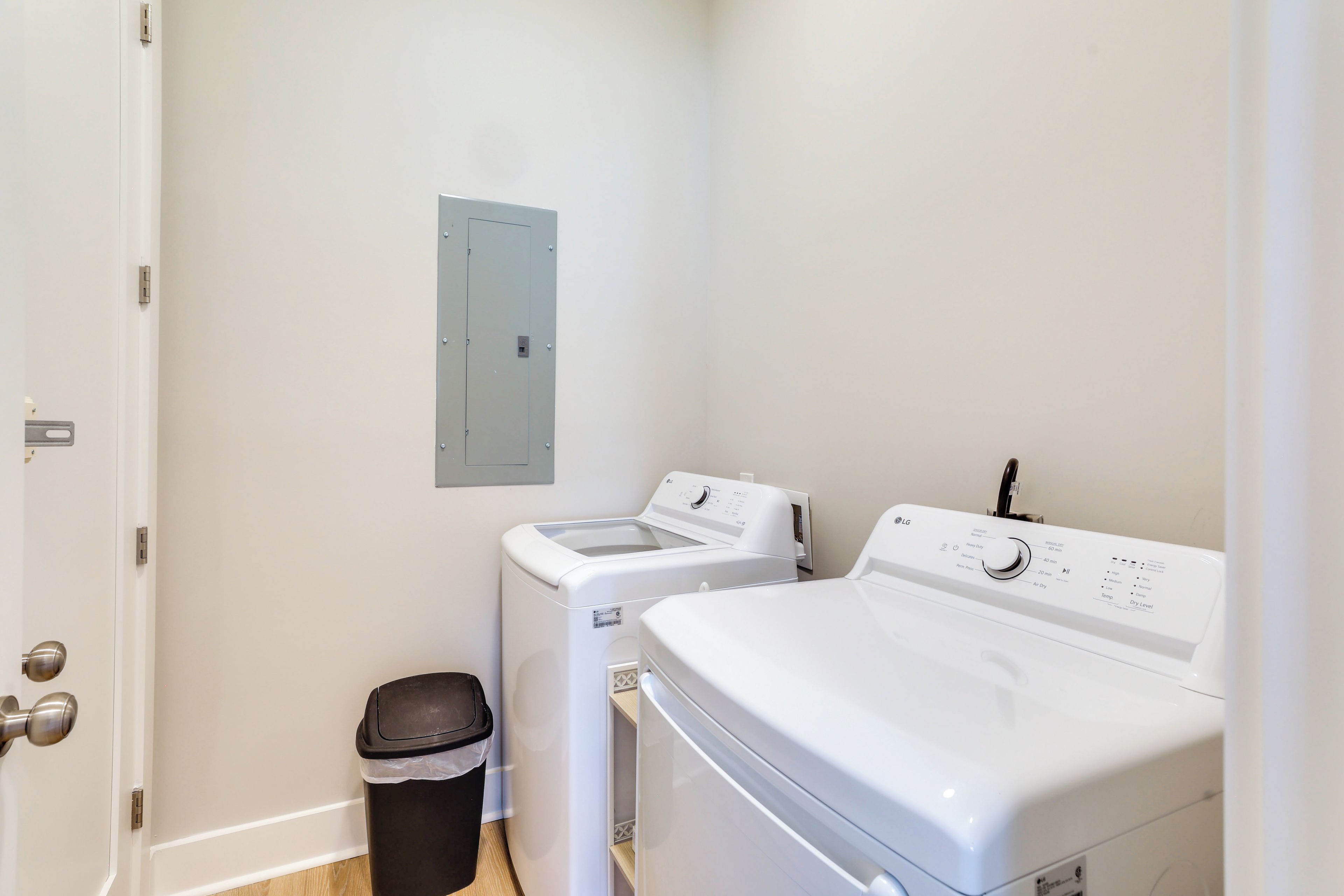 A laundry room with a front-loading washer and dryer, a trash bin, and a utility panel on the wall.