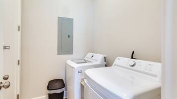 A laundry room with a front-loading washer and dryer, a trash bin, and a utility panel on the wall.