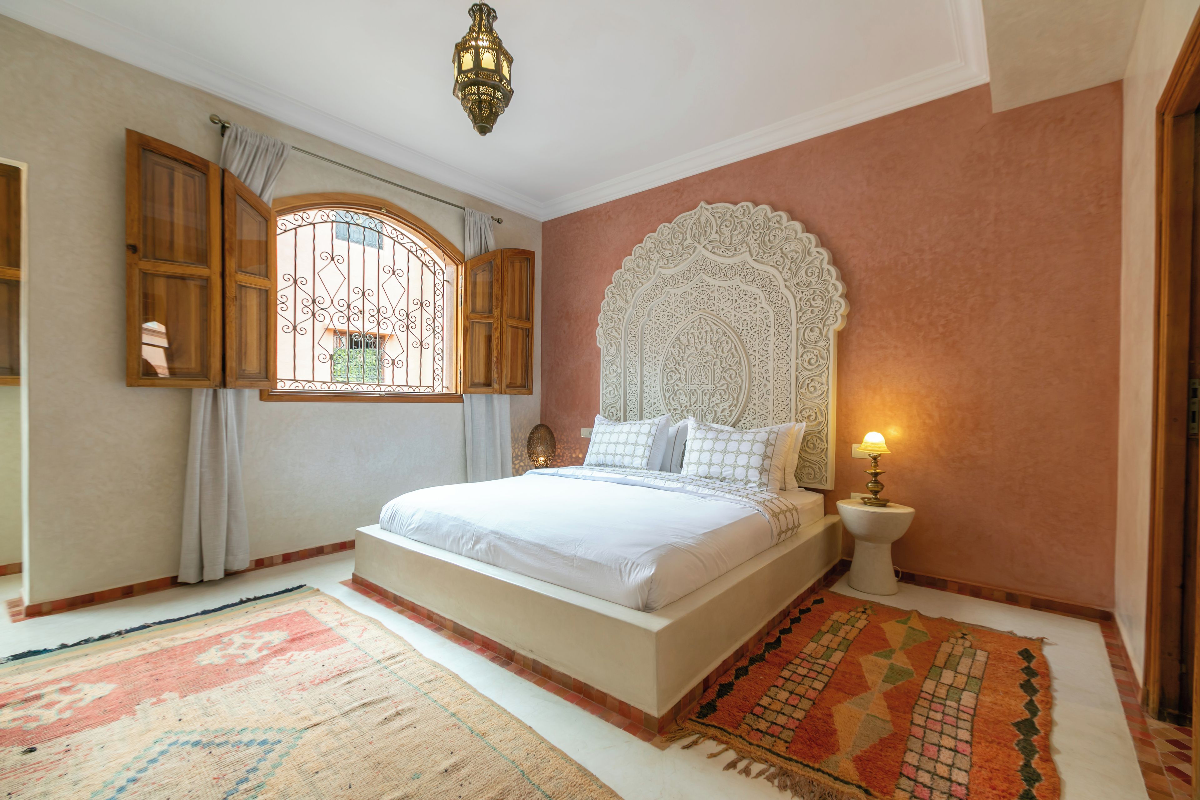 Een slaapkamer met een groot bed, een decoratief hoofdeinde, een nachtkastje met een lamp en een raam met gordijnen.