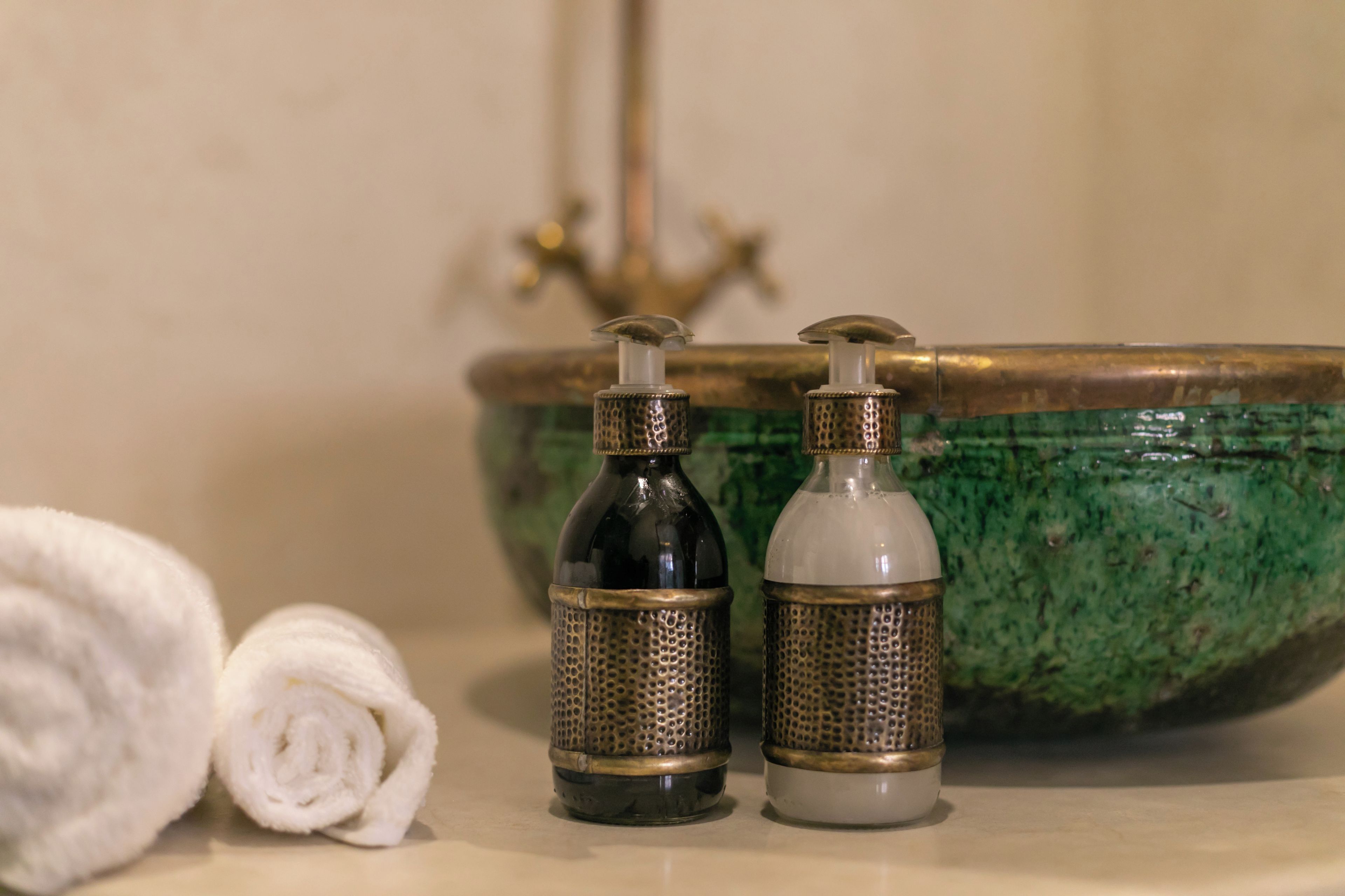 Dos botellas metálicas con dosificadores tipo bomba, una toalla blanca y un lavamanos verde con grifo dorado en un baño.