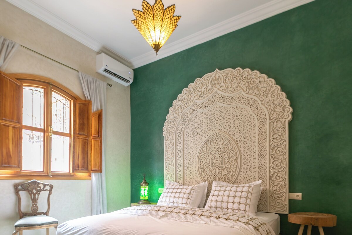 A bedroom with a large bed, a decorative headboard, a chandelier, a window with curtains, and a patterned rug.