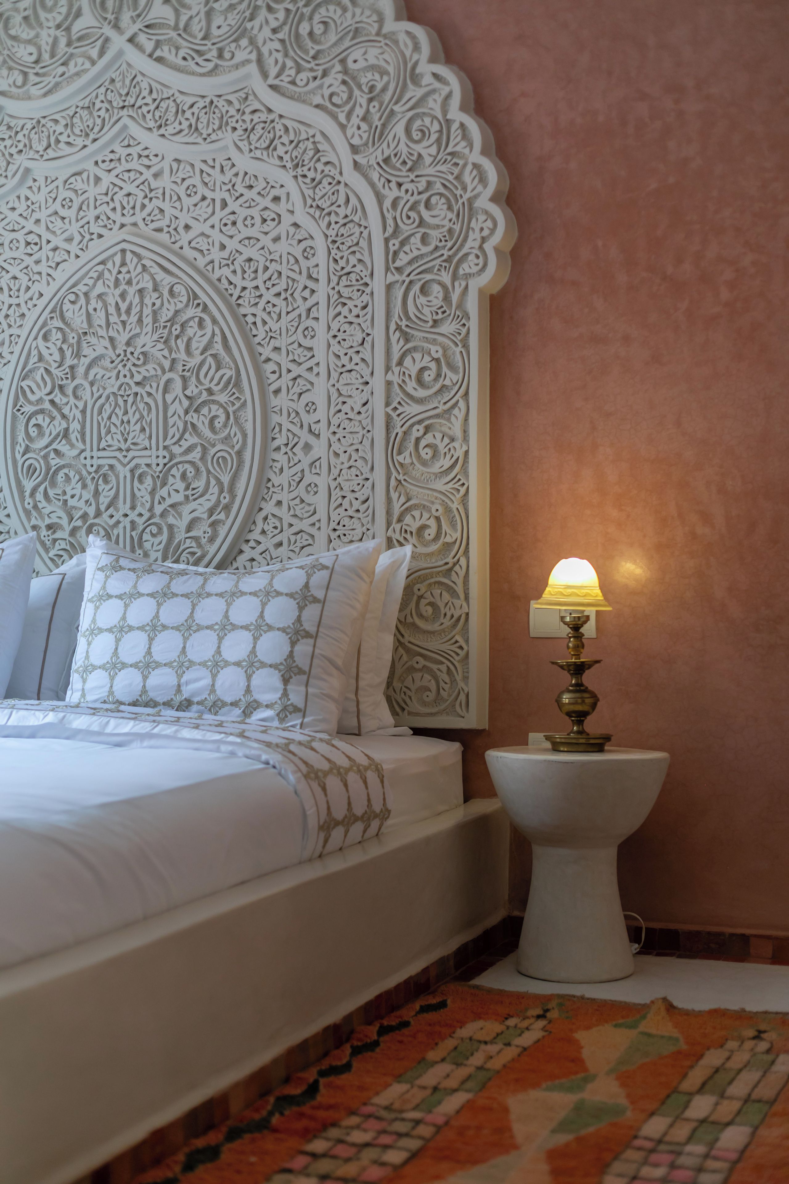 Una cama con cabecera blanca y almohadas estampadas, una lámpara de noche y una alfombra con diseño.