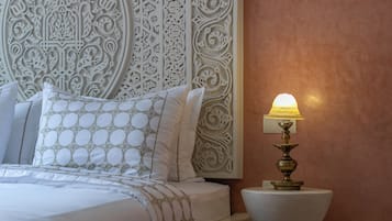 Una cama con cabecera blanca y almohadas estampadas, una lámpara de noche y una alfombra con diseño.