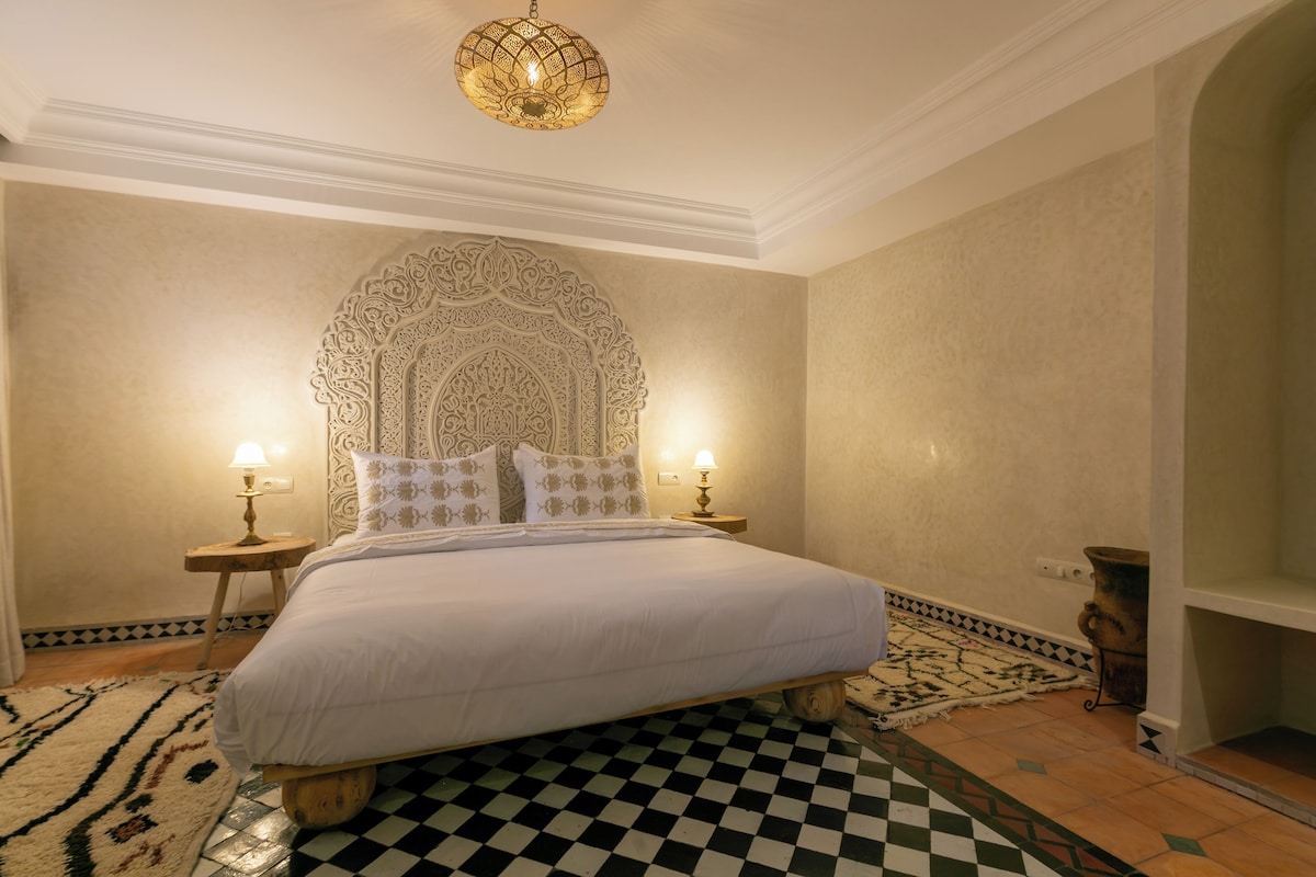 A hotel room with a large bed, ornate headboard, two bedside lamps, and a patterned rug.