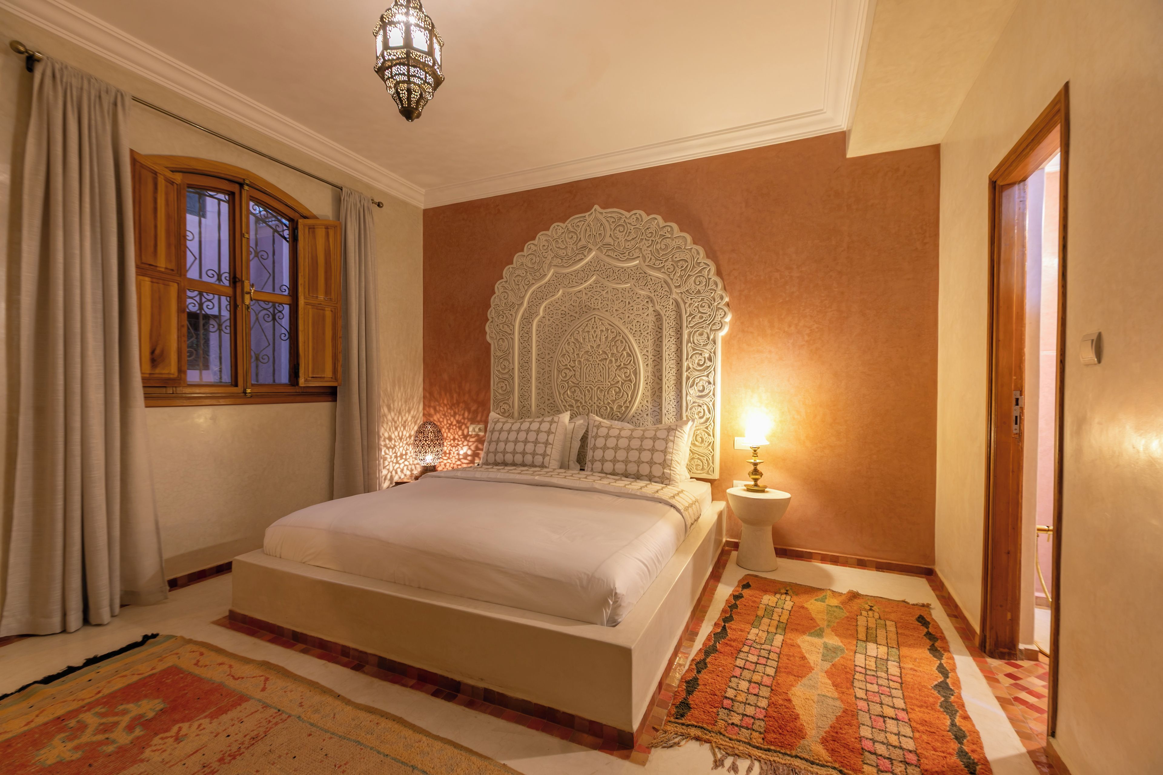 Een slaapkamer met een groot bed, een decoratief hoofdbord, een nachtkastje met een lamp en een kamerkleed met een patroon.
