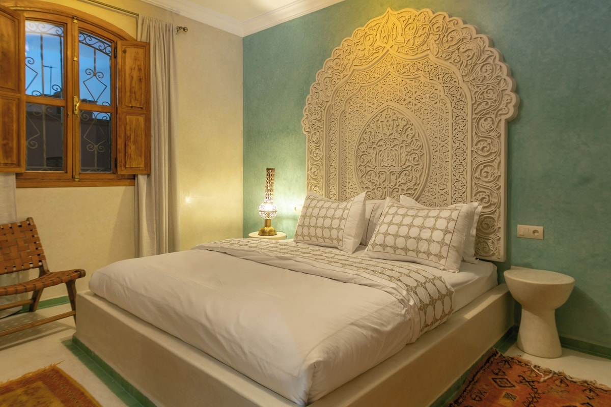 A bedroom with a large bed, a decorative headboard, a nightstand, a lamp, a chair, and a patterned rug.
