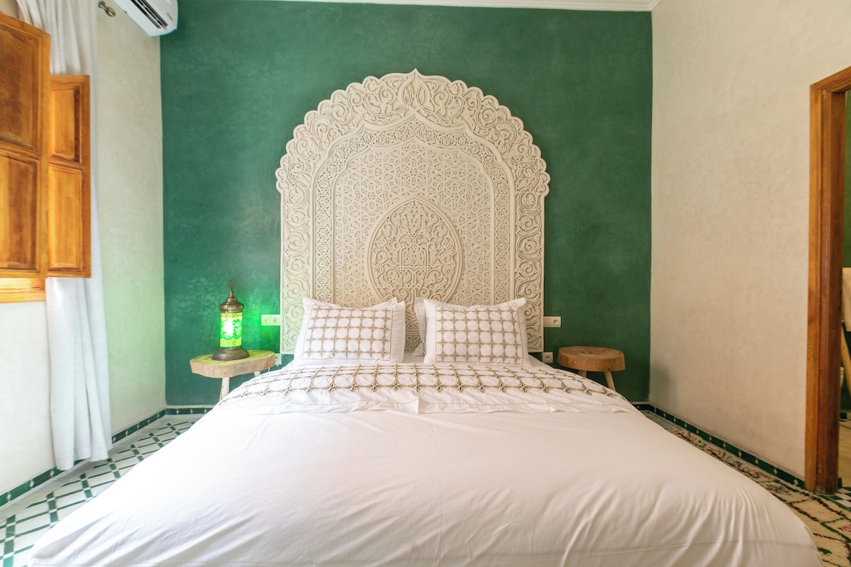A bed with a white bedspread and pillows, a green wall, and a decorative headboard.