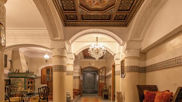 Un pasillo ornamentado con alfombra estampada, un candelabro y una vista hacia un cuarto con piso a cuadros.