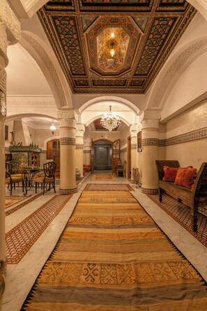 An ornate hallway with a patterned carpet, a chandelier, and a view into a room with a patterned floor. - Riad Dar Razallah  (Marrakech)