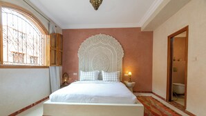 Ein Schlafzimmer mit einem großen Bett, einem verzierten Kopfteil, einem Fenster mit Metallgitter, einer Nachttischlampe und einem durch eine offene Tür einsehbaren Badezimmer.