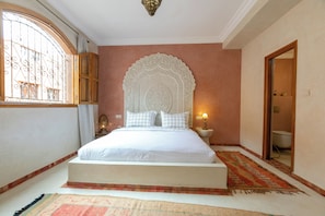 A bedroom with a large bed, ornate headboard, a window with metal bars, a bedside lamp, and a bathroom visible through an open door. - Riad Dar Razallah  (Marrakech)