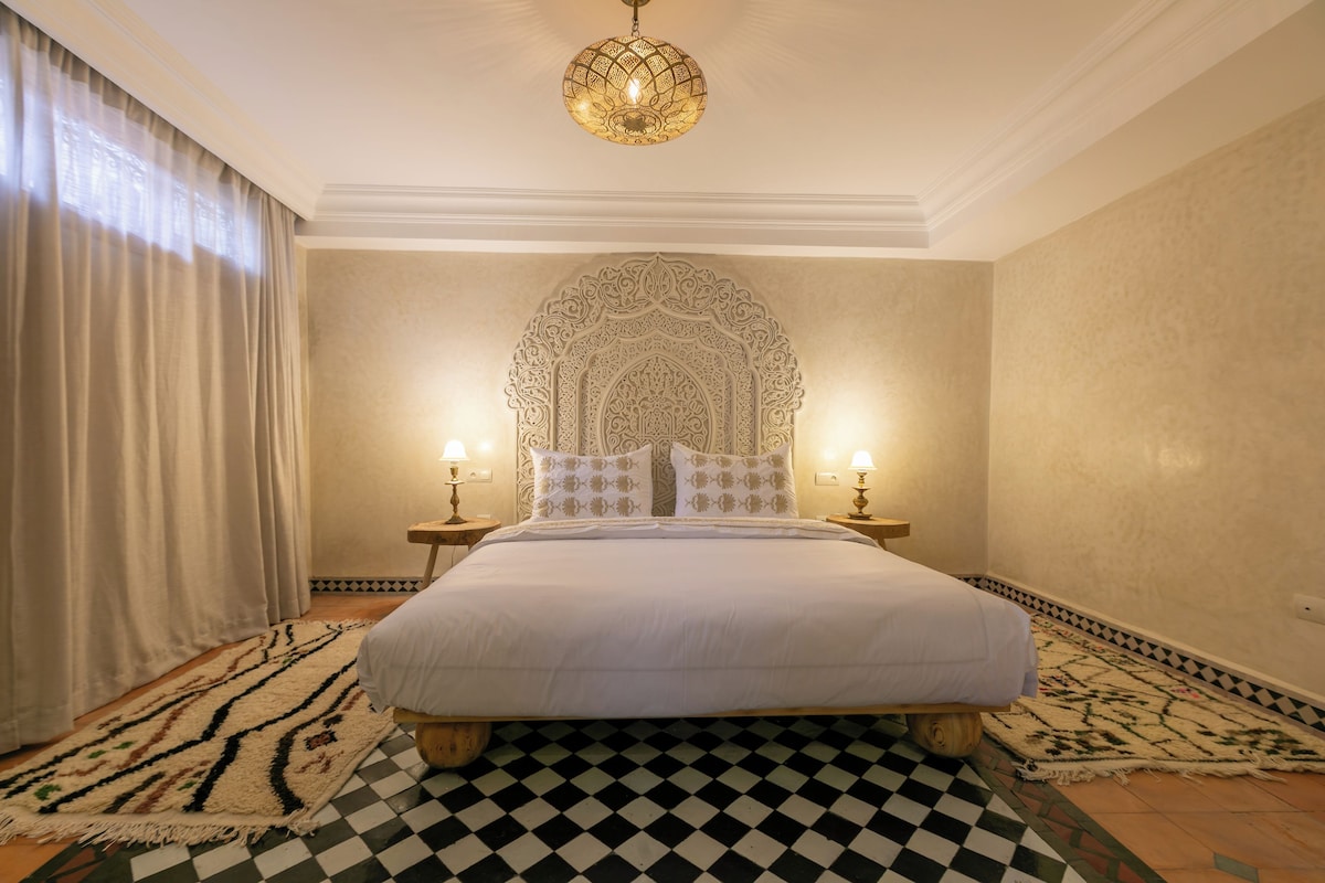 Una habitación de hotel con una cama grande, dos lámparas de noche, un tapete estampado y una lámpara decorativa en el techo.