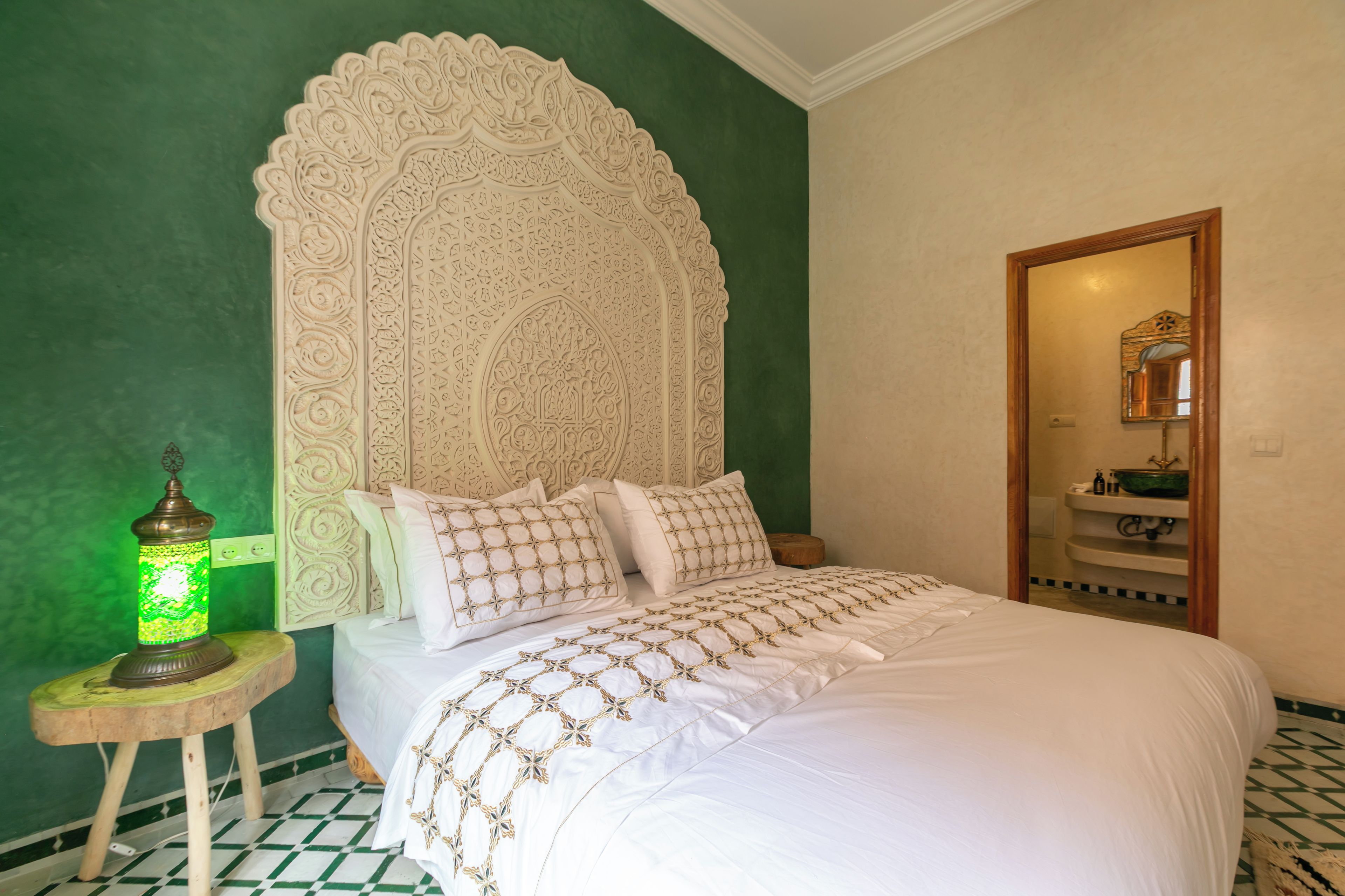 Un dormitorio con un gran cabecero ornamentado, una cama con ropa de cama blanca y bordados dorados, una pared verde, un taburete pequeño con una lámpara verde, y una puerta de madera que da al baño.