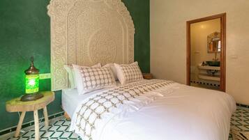 Un dormitorio con un gran cabecero ornamentado, una cama con ropa de cama blanca y bordados dorados, una pared verde, un taburete pequeño con una lámpara verde, y una puerta de madera que da al baño.