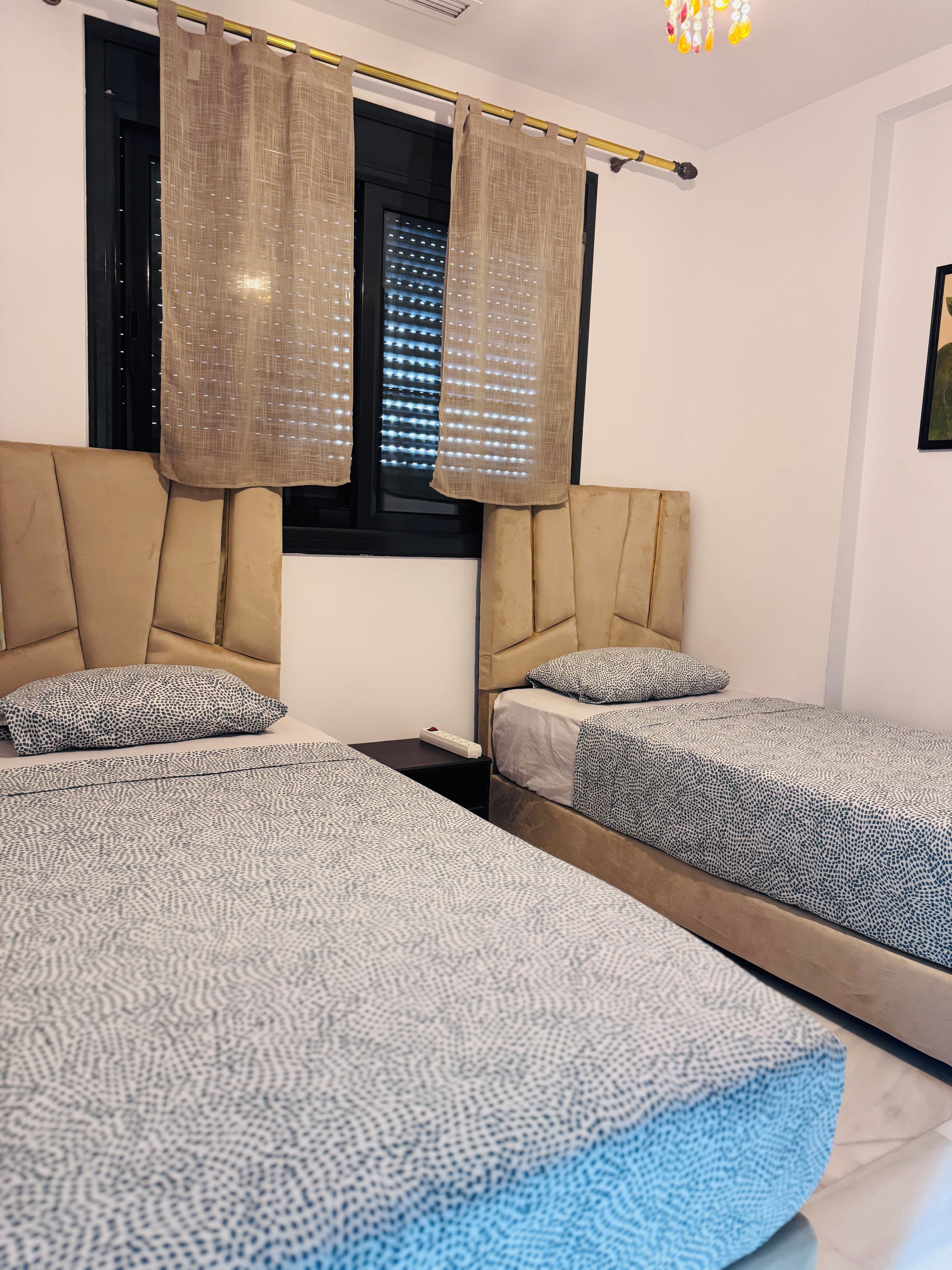 A hotel room with two beds, a nightstand, a window with blinds, and a ceiling light fixture.