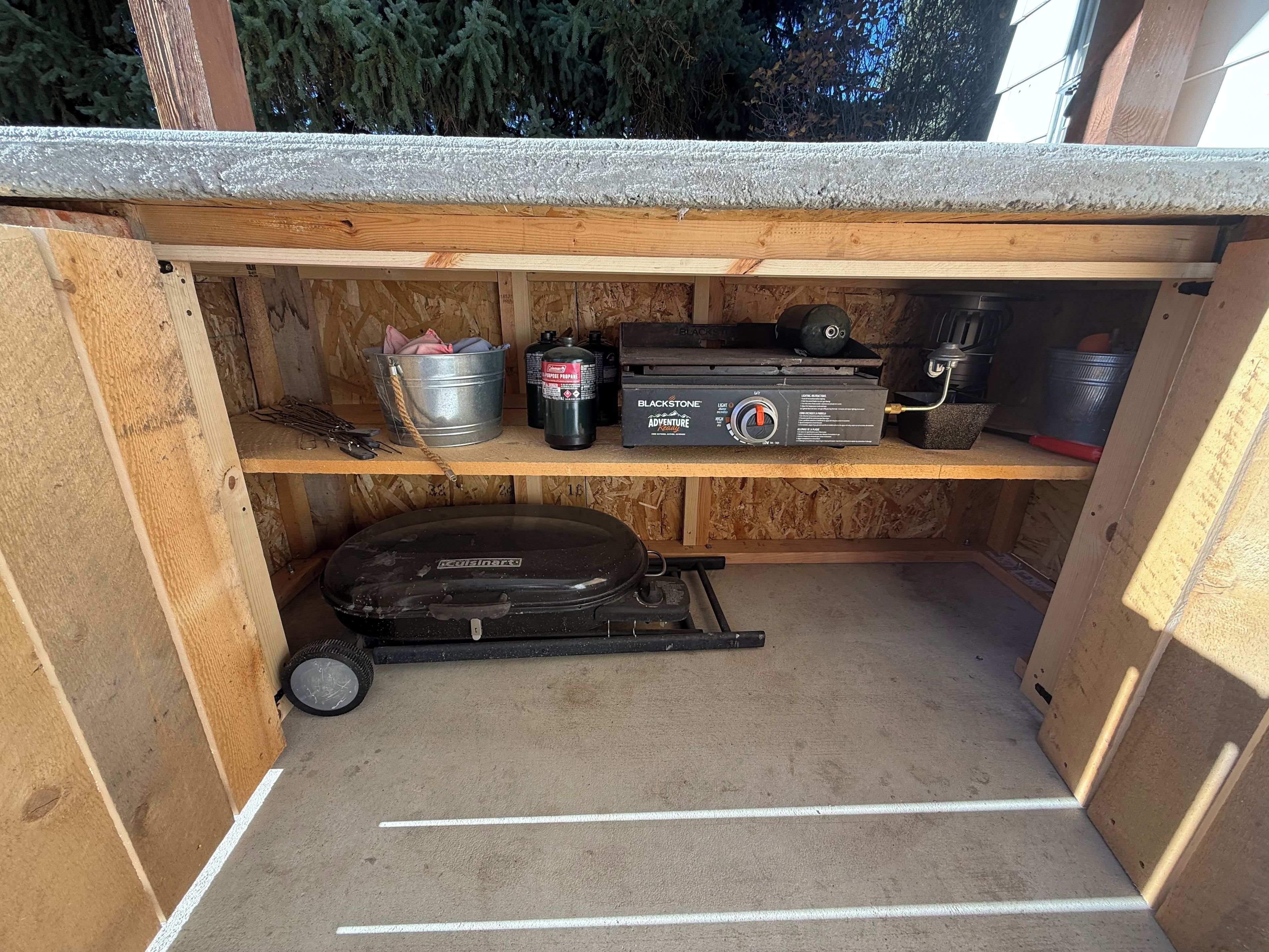 Ein kleiner, abgeschlossener Raum mit Betonboden, Holzvertäfelung und einem Regal mit verschiedenen Gegenständen, darunter ein Grill, ein Kanister und eine Kiste mit dem Aufdruck 'BLACKSTONE Adventure'.