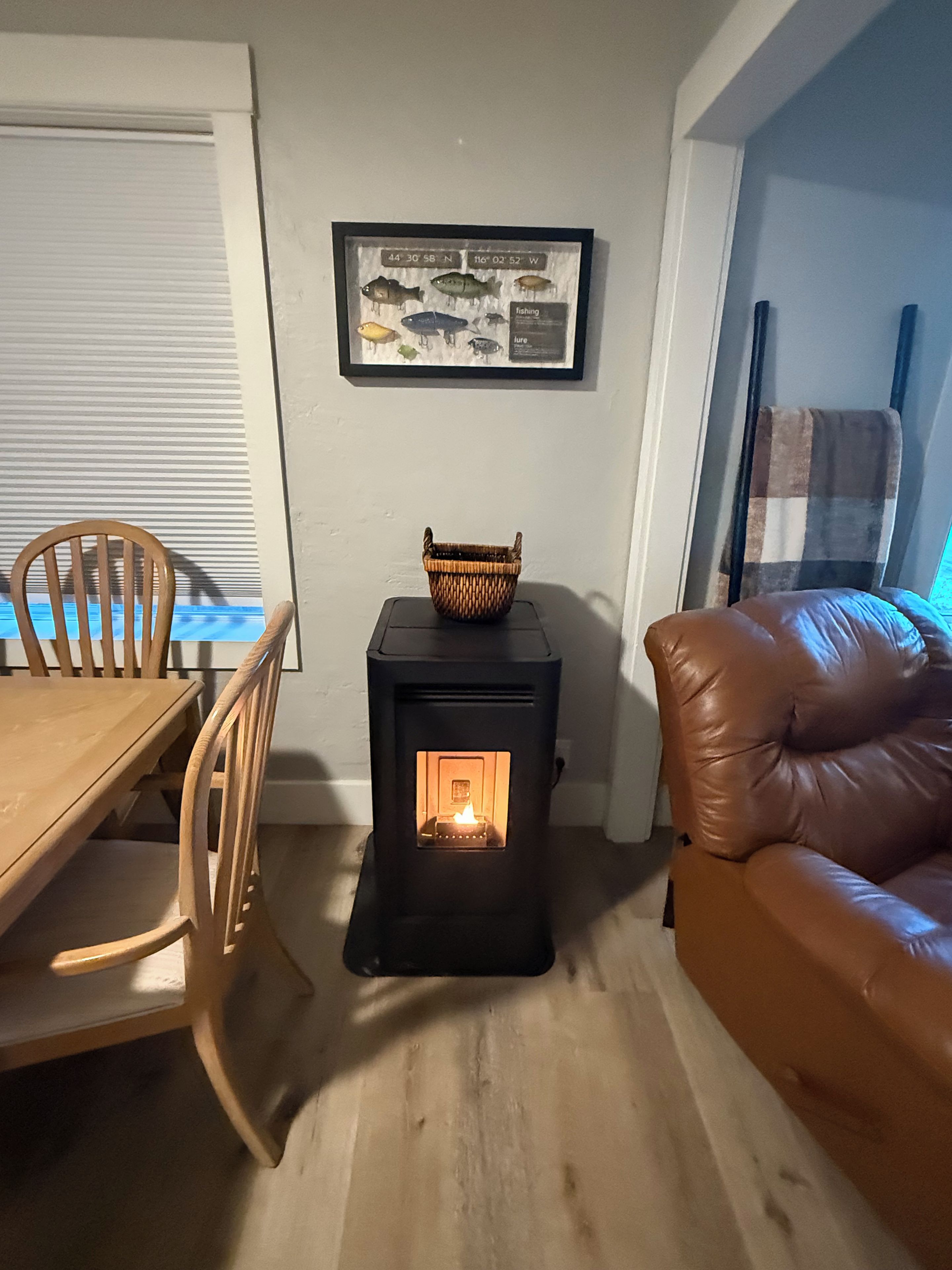 A room with a wooden dining table, a brown leather sofa, a black wood-burning stove, and a framed fish illustration on the wall.