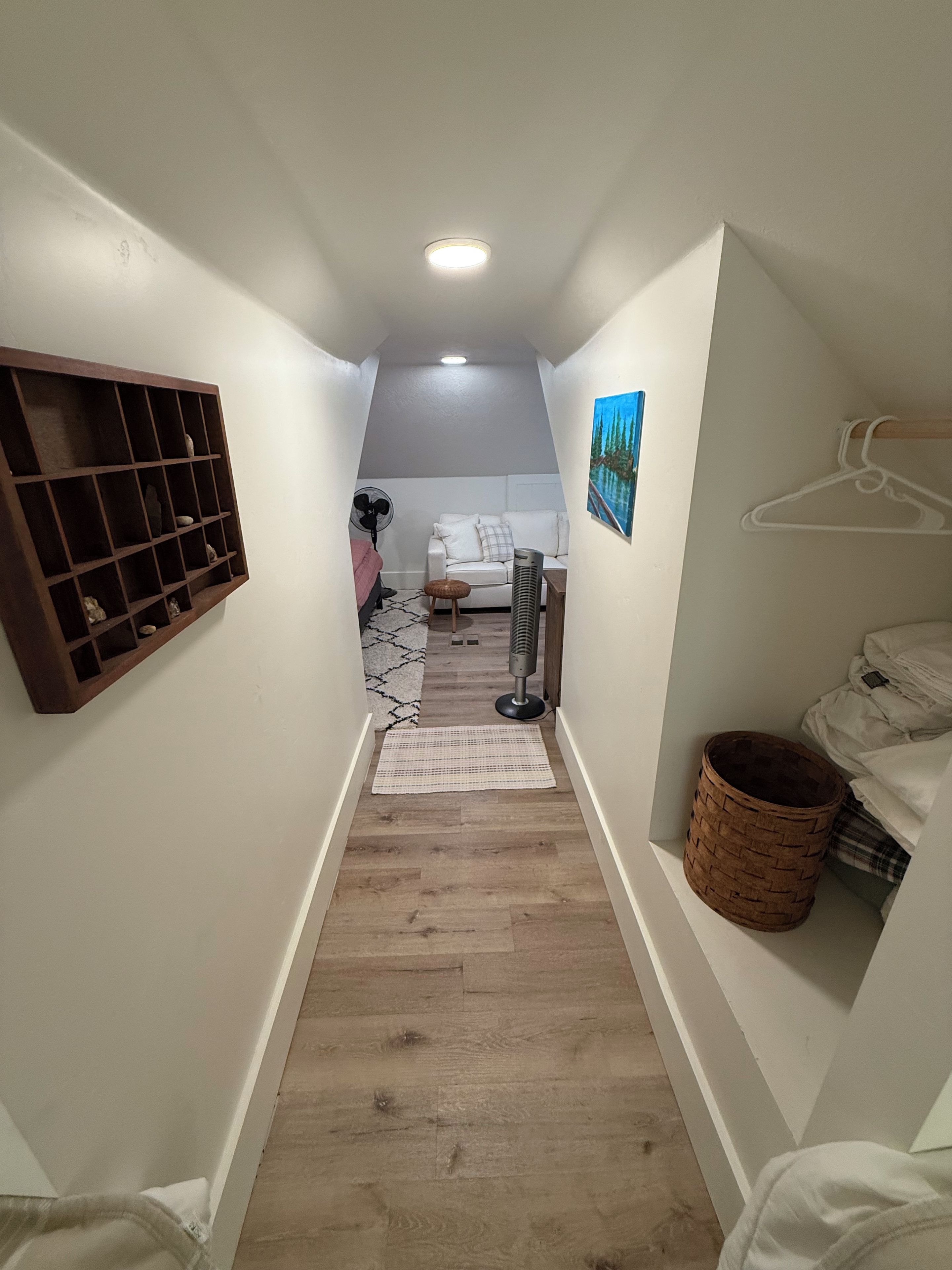 A narrow hallway with a wooden floor, a white wall with a shelf, a white couch, a fan, and a painting.