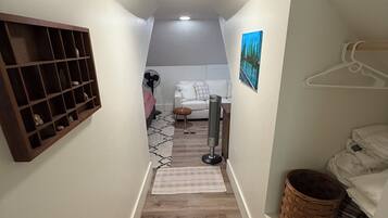 A narrow hallway with a wooden floor, a white wall with a shelf, a white couch, a fan, and a painting.