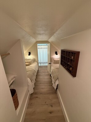 A narrow, sloped hallway with two beds on either side, a wooden floor, and a window at the end.
