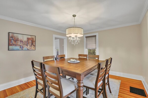 A dining room with a wooden table and chairs, a chandelier, a painting on the wall, and a window with blinds.