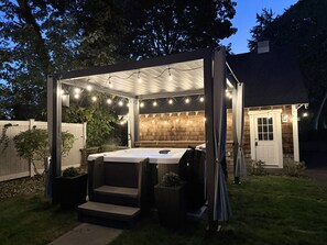 A hot tub under a covered patio with string lights, surrounded by a wooden fence and a house.