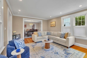 A living room with a beige sofa, a blue armchair, a coffee table, and a dining area in the background. - Serenity Retreat | Hot Tub + Fire Pit | Walk Downtown (West Hartford)