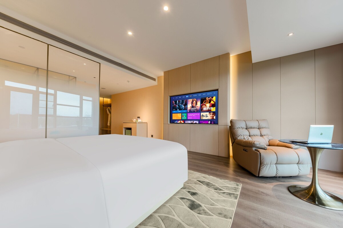 Ein modernes Schlafzimmer mit einem großen Bett, einem an der Wand montierten Fernseher, einem Ledersessel und einem kleinen Tisch mit einem Laptop.