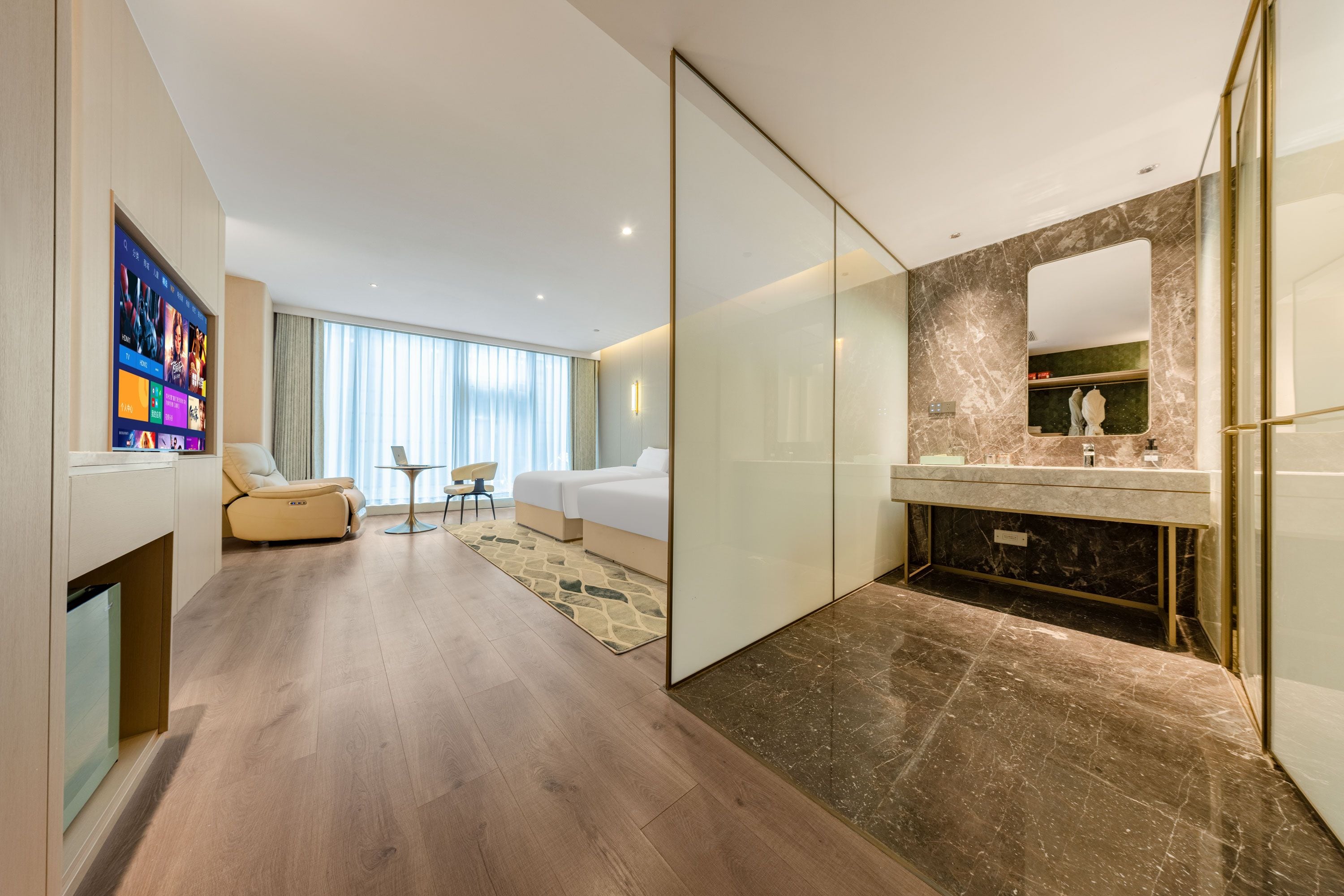 A modern hotel room with a large bed, a separate seating area, a bathroom with a marble vanity, and a flat-screen TV mounted on the wall.