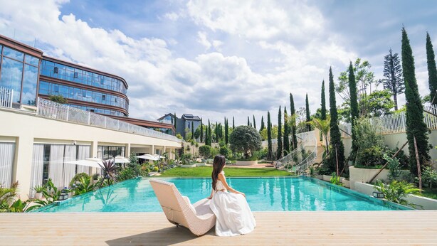 A poolside area with a person sitting on a lounge chair, a pool, and a modern building in the background. - SKANDURA GARDEN HOTEL DIANCHI (Kunming)