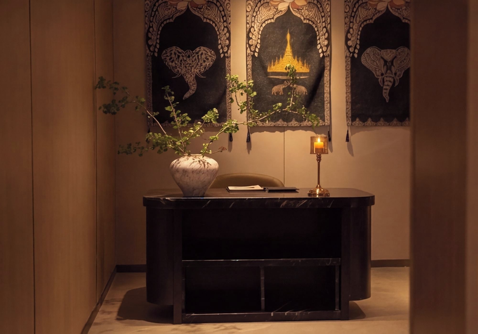 A dark wooden desk with a potted plant, a lit candle, and a chair in a warmly lit room with decorative wall art.