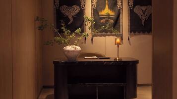 A dark wooden desk with a potted plant, a lit candle, and a chair in a warmly lit room with decorative wall art.