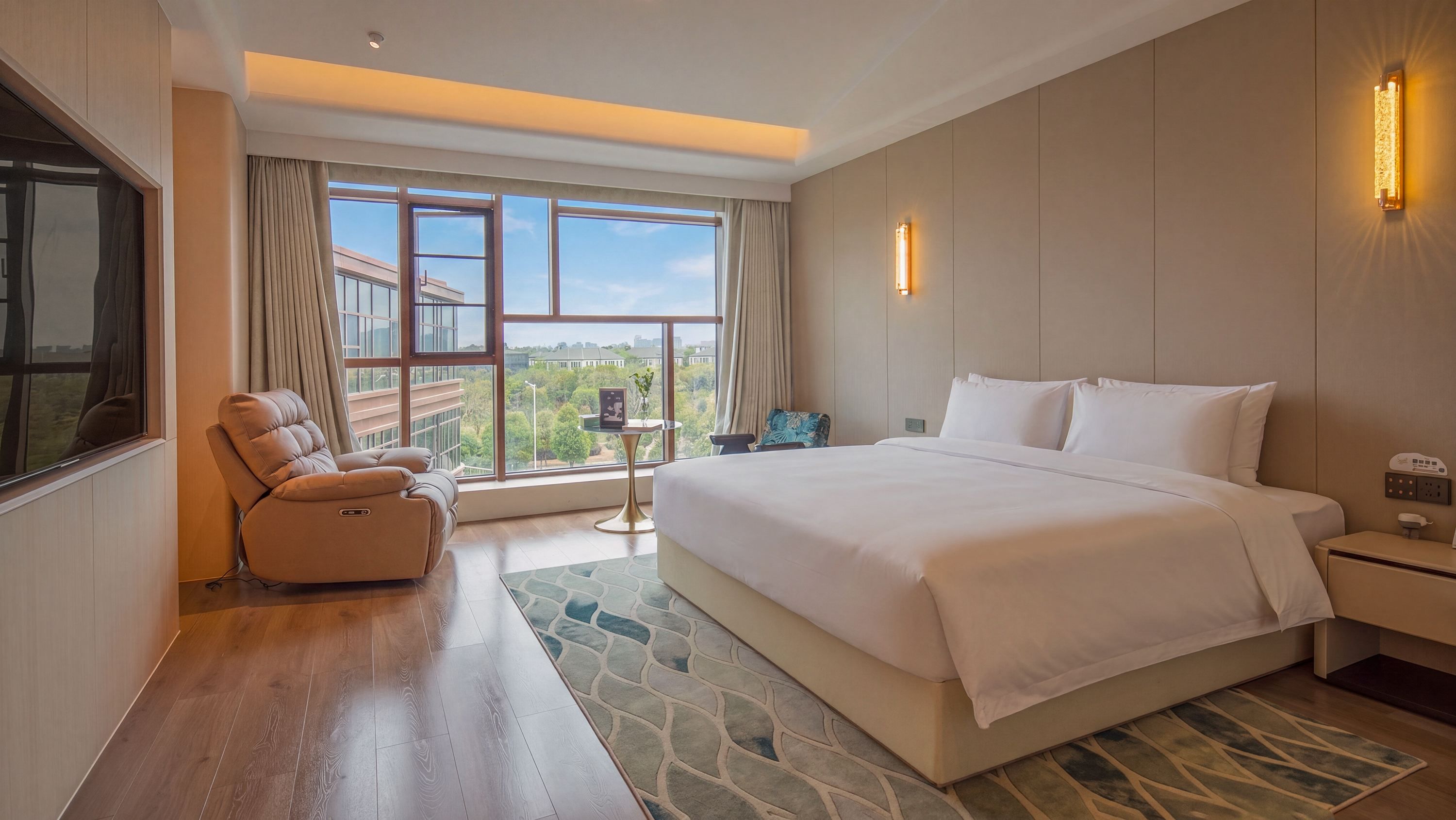 A hotel room with a large bed, a seating area with a chair, a view of the outdoors through large windows, and a wall-mounted light fixture.