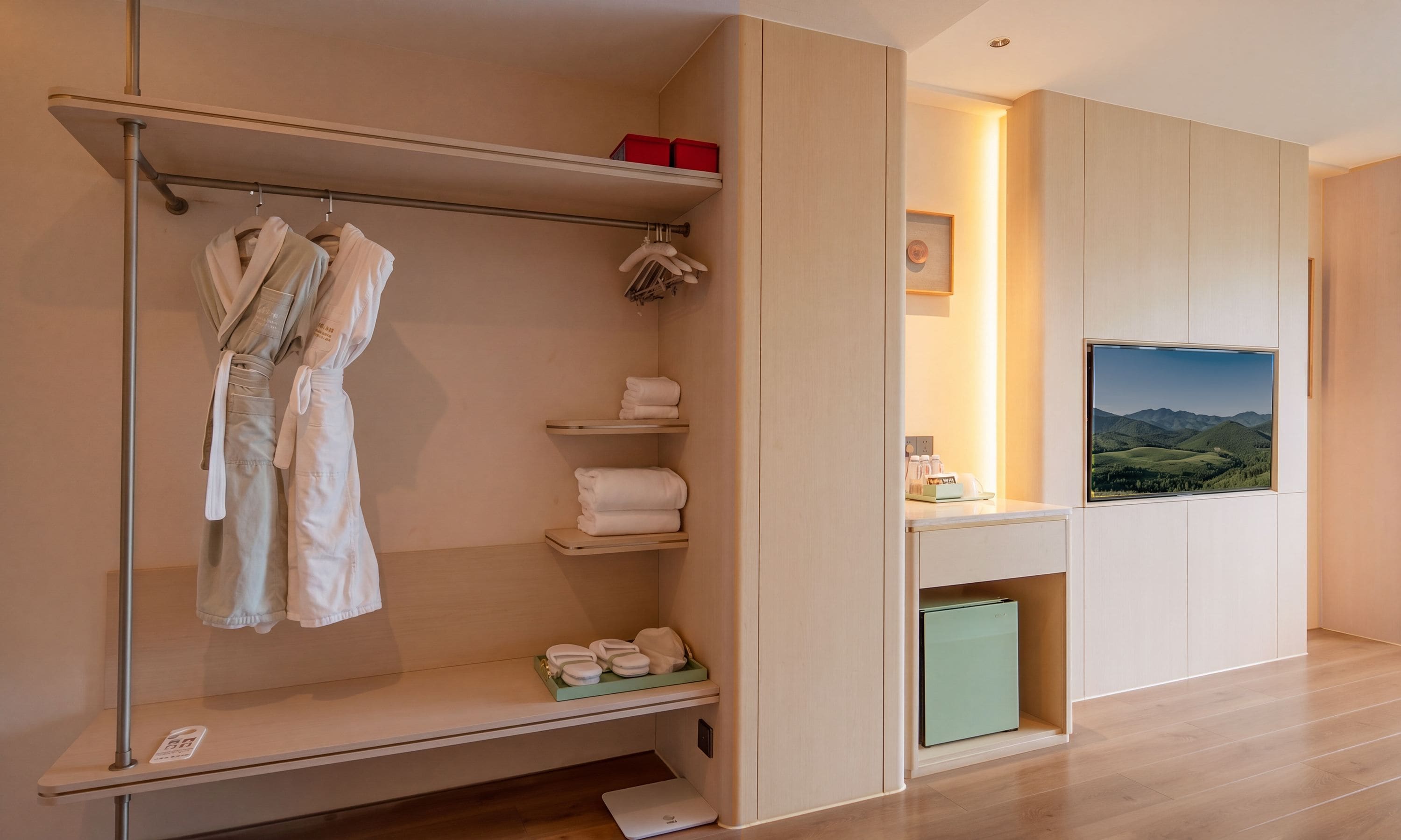 A hotel room with a robe and towel rack, a small shelf with towels, a mini fridge, and a view of the outdoors.