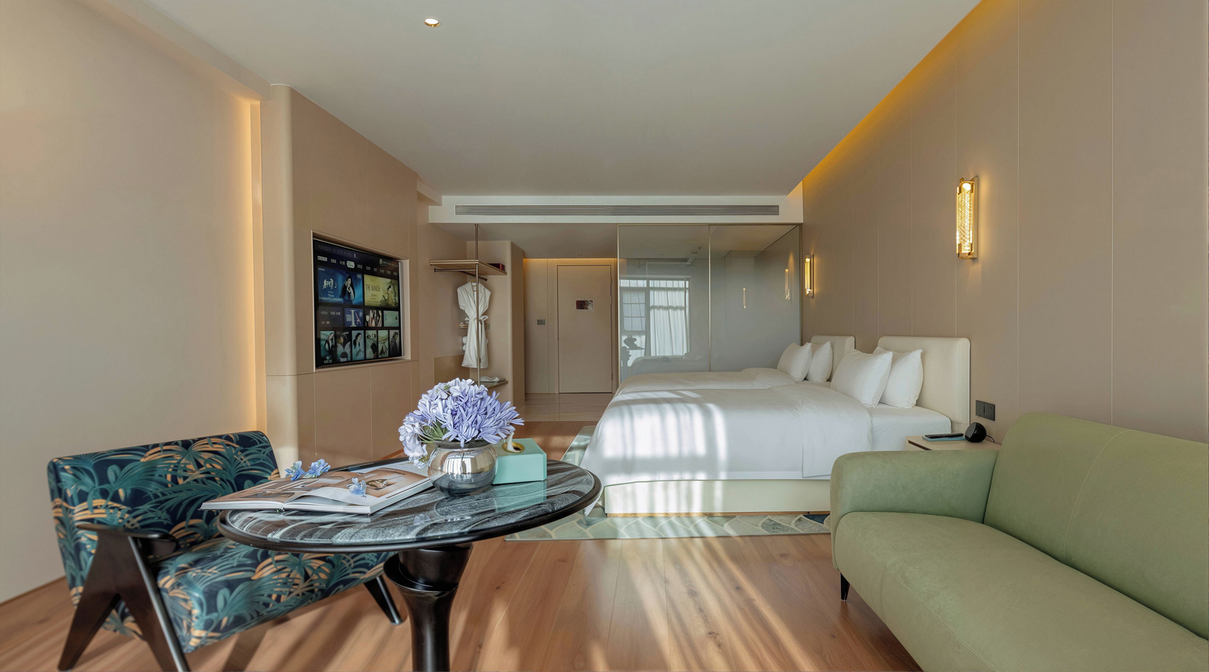 Una habitación de hotel con cama, sofá verde, una mesa redonda con un florero, una silla y televisor montado en la pared.