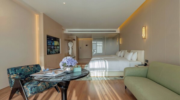 A hotel room with a bed, a green sofa, a round table with a vase, a chair, and a wall-mounted TV.